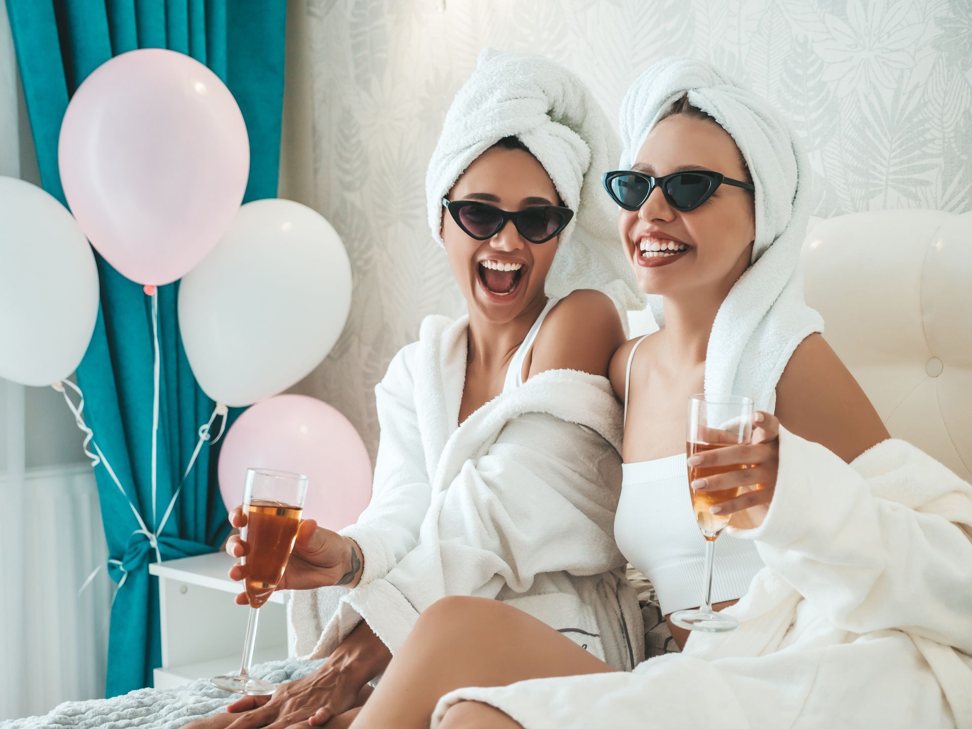 Two women in bathrobes and towels on their heads are sitting on a bed holding wine glasses.