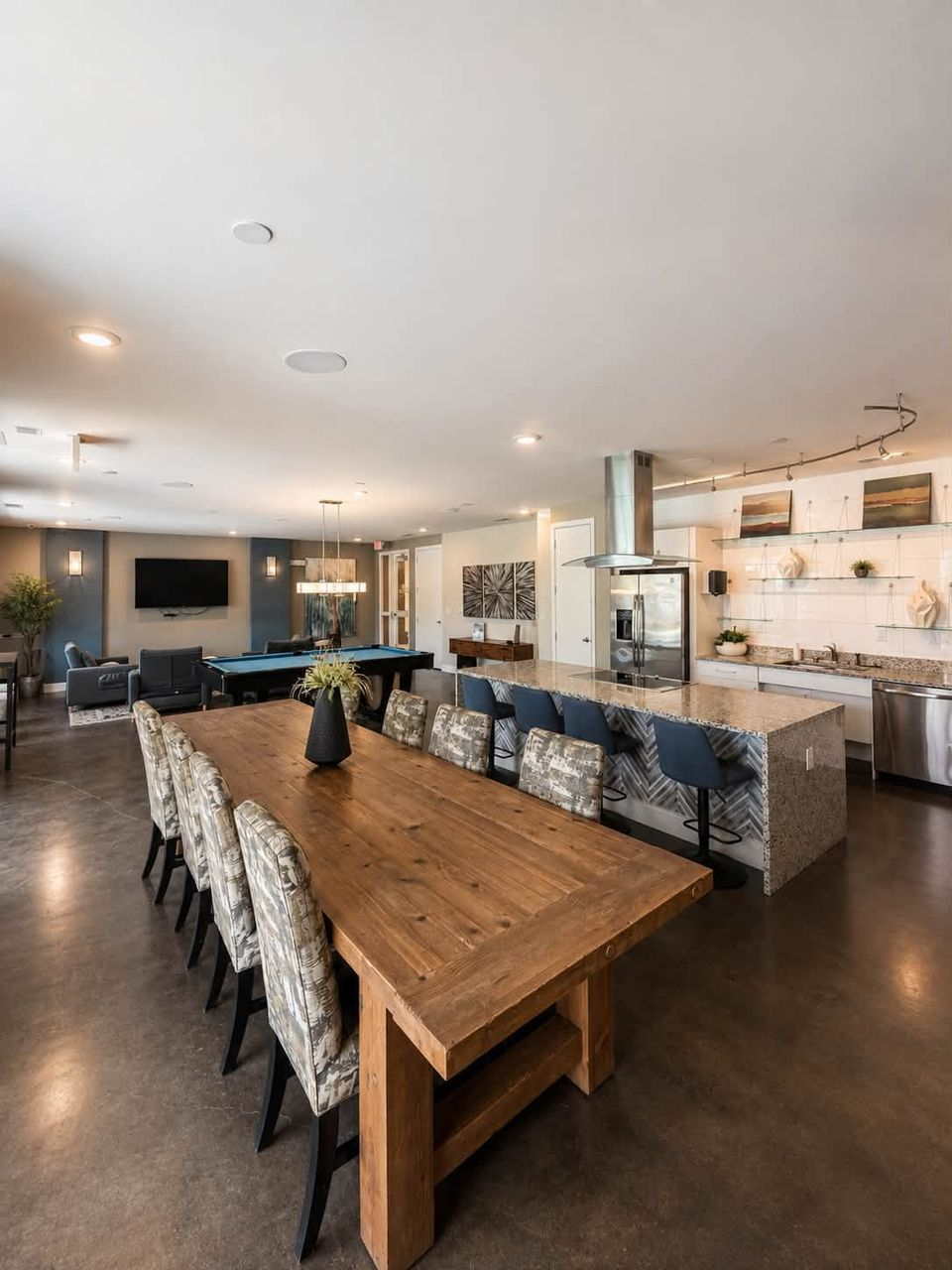 Modern club room and kitchen with a long wooden dining table and patterned chairs.