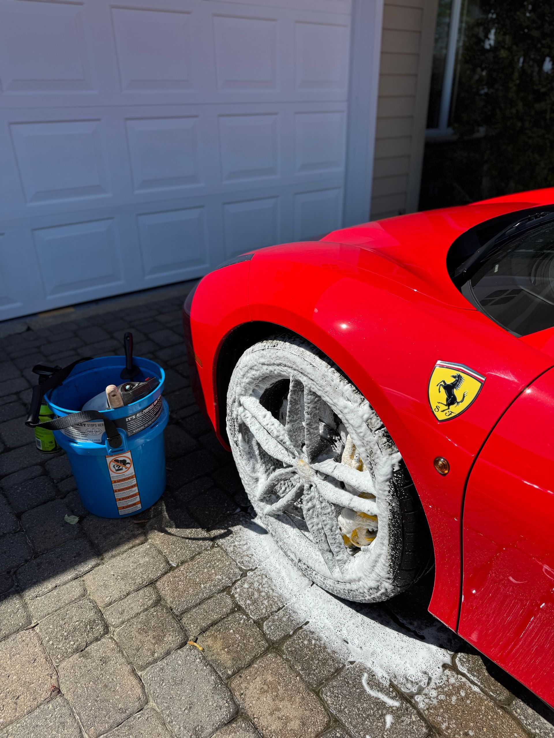A red Ferrari being washed, with foamy soap on the wheel and a bucket of supplies nearby, in front of a garage.