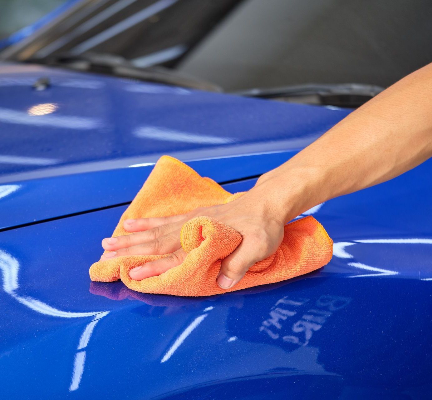 Hand wiping a bright orange microfiber cloth across the blue hood of a car, cleaning and polishing the surface.