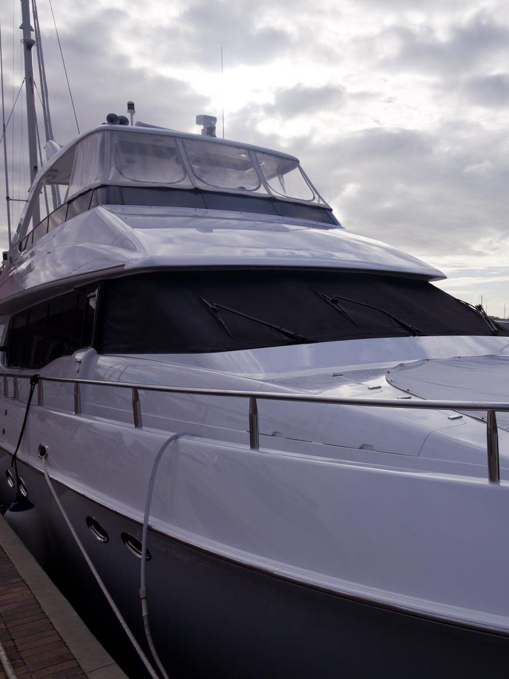 White motor yacht docked at a pier, with a gray hull and black covers over the front windows, under a cloudy sky.