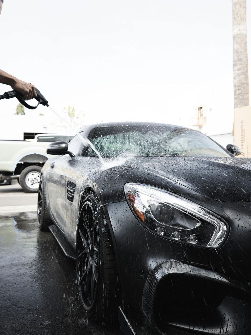 A person washes a matte black sports car with a hose. The car is wet with water.