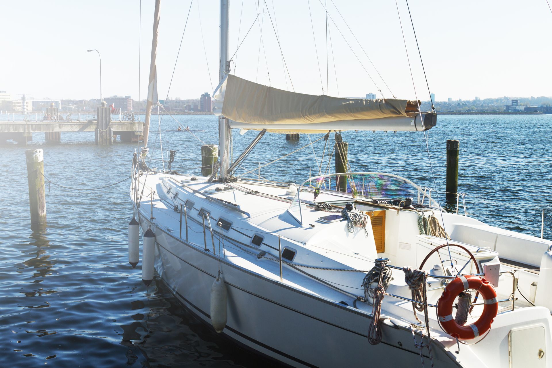 White sailboat docked at a pier on a sunny day, with a beige sail unfurled. Calm blue water.