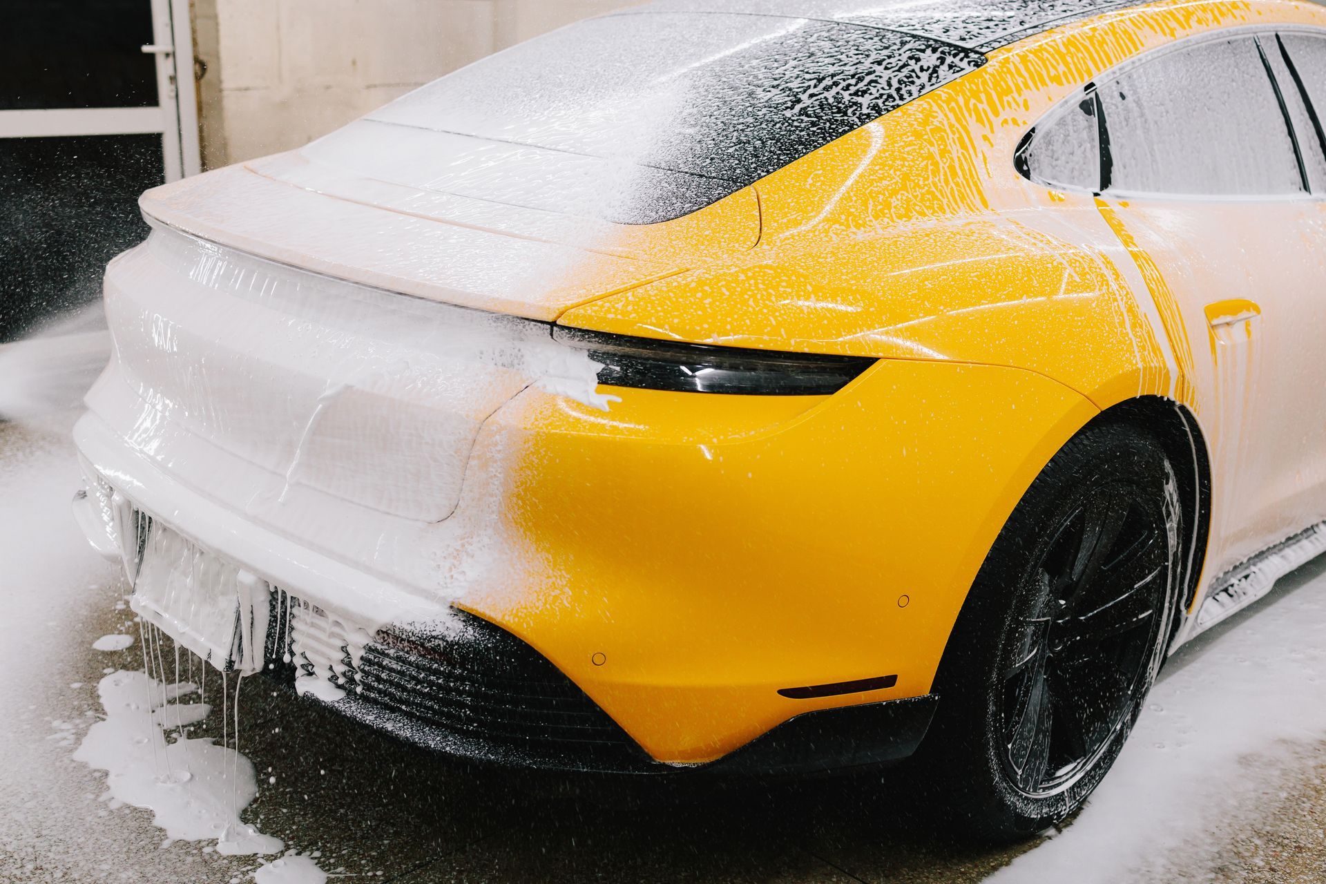 A yellow Porsche Taycan being washed with white foam in a carwash. The rear of the car is covered in suds.
