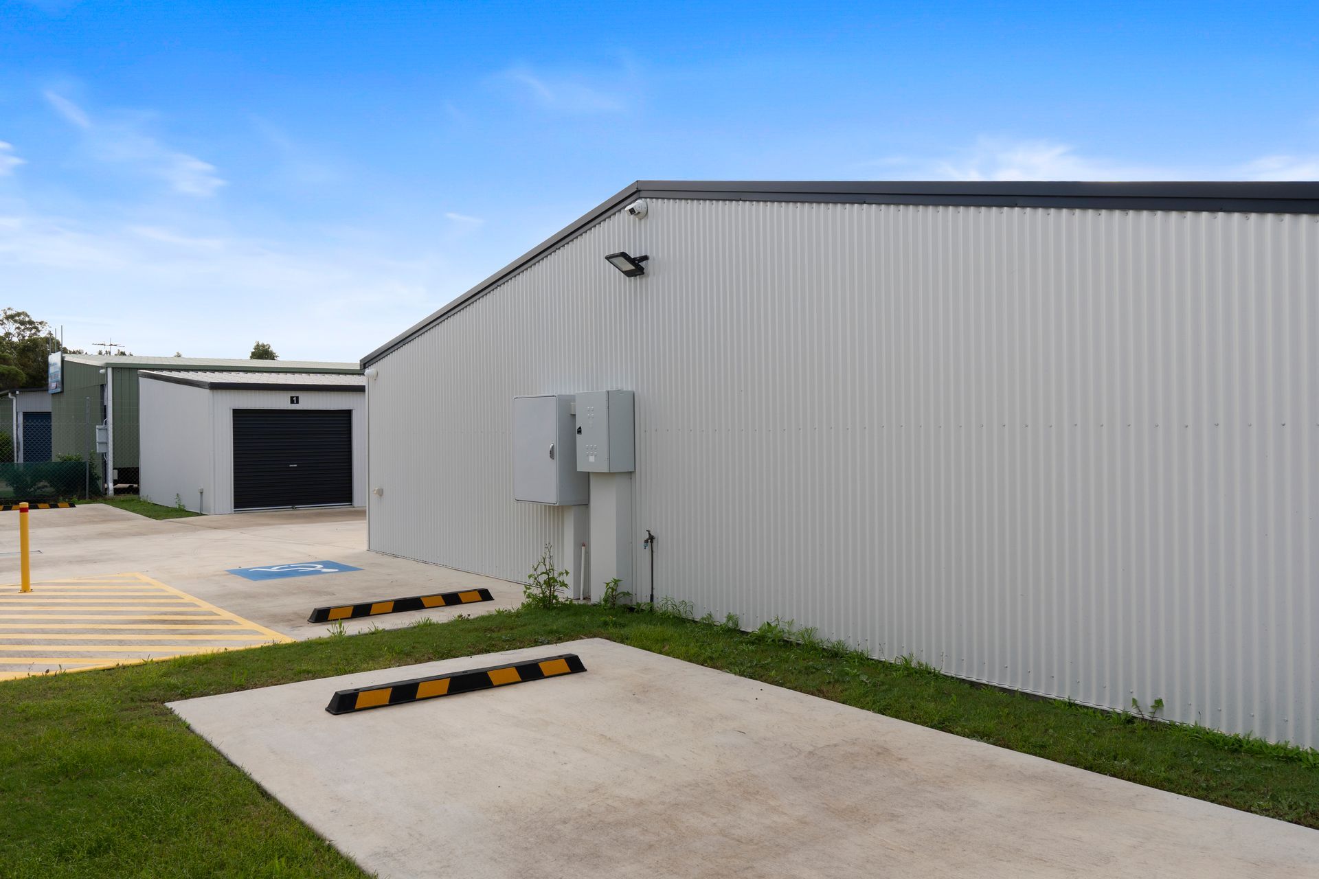 Exterior view of a storage facility with multiple units. Gray buildings, black doors, concrete ground, blue sky.