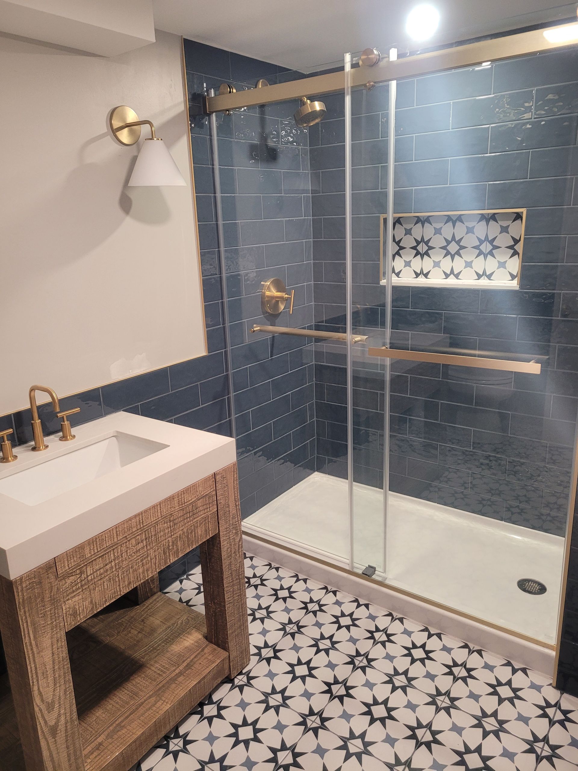 Bathroom with blue tile shower, patterned floor, wood vanity, gold fixtures, and a glass shower door.