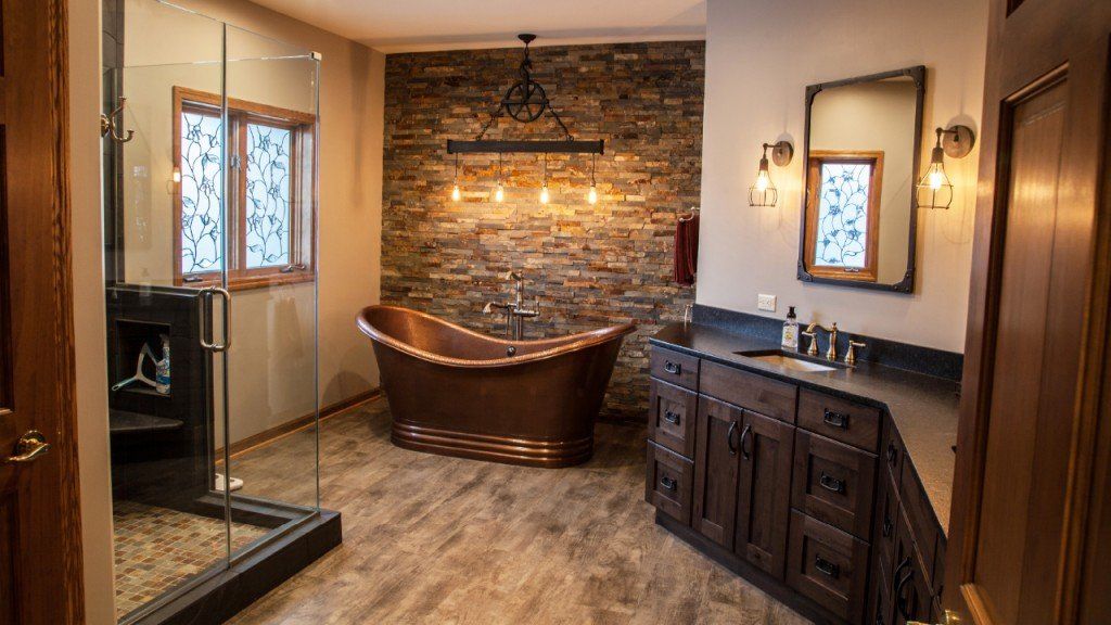 Bathroom with copper bathtub against a stone wall, glass shower, wooden vanity, and tiled floor.
