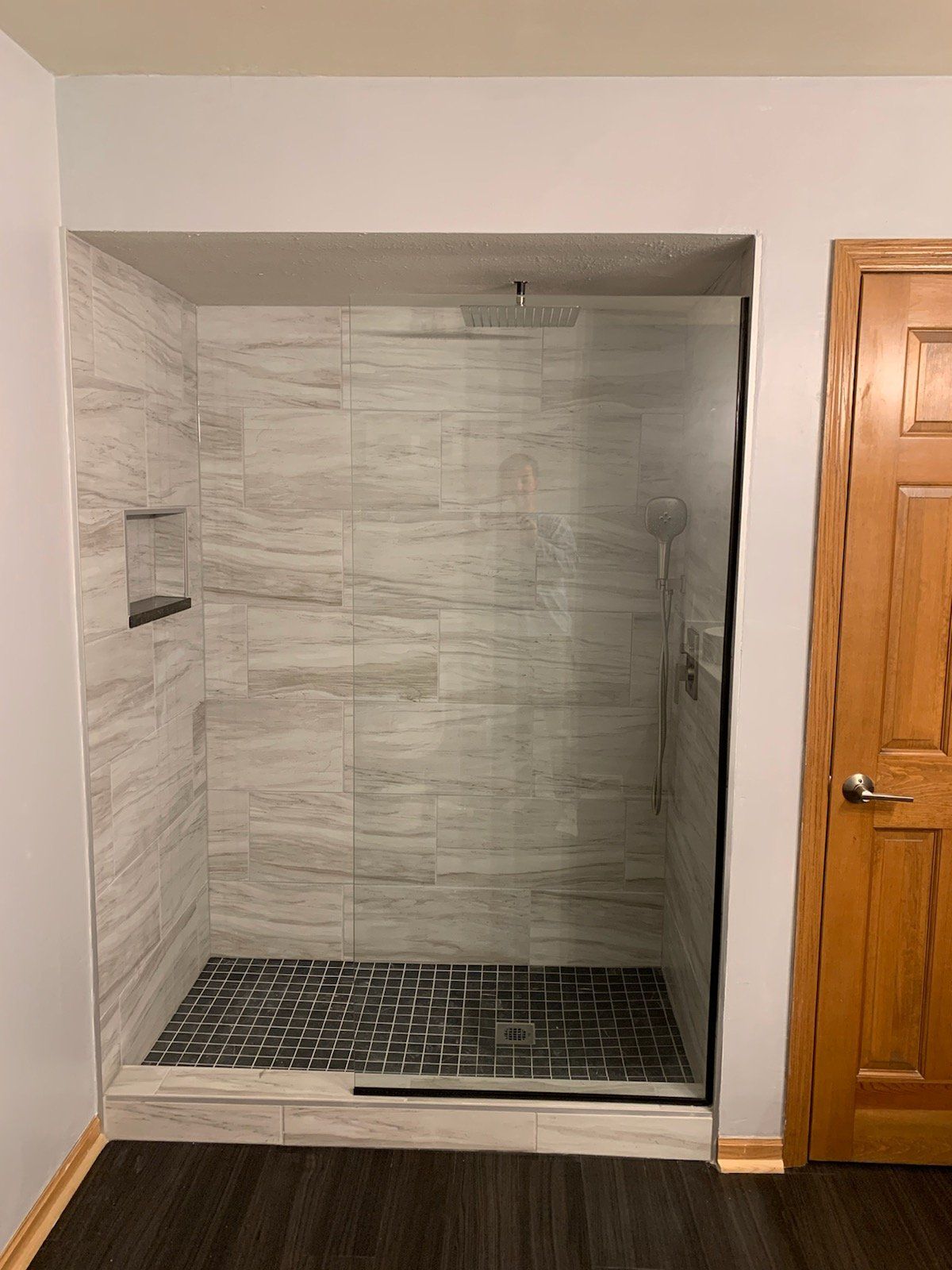 A newly renovated shower with gray-toned tile and dark floor, framed by a glass door, next to a wooden door.