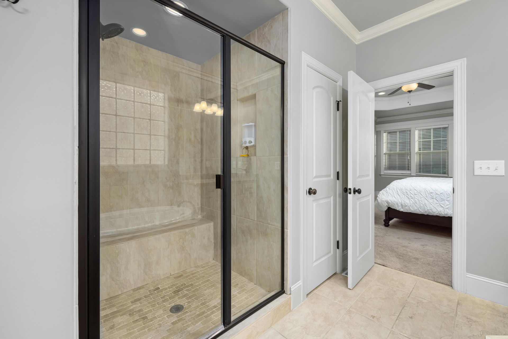 Bathroom with a glass shower enclosure, beige tile, and an open doorway to a bedroom.