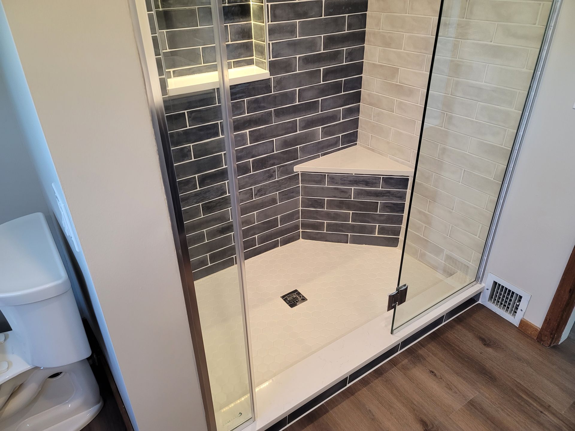 Modern corner shower with glass doors, white tiled base, and gray brick-style wall tile with a built-in bench.
