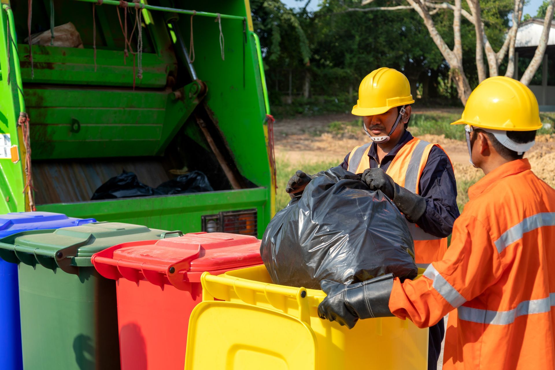 Two men wearing hard hats are loading garbage into a garbage truck.