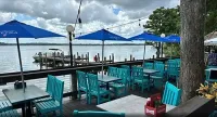 Outdoor waterfront restaurant seating with turquoise chairs, blue umbrellas, and cloudy sky over the water