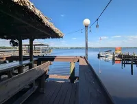 Sunlit dockside deck with benches, thatched roof, and calm blue water with small boats