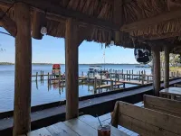 Thatched-roof lakeside dock with wooden posts, calm water, and a red chair in the distance