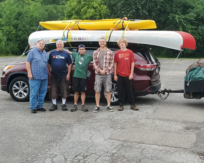 two middle age men and three teens standing in front of a car with canoes strapped to the top.