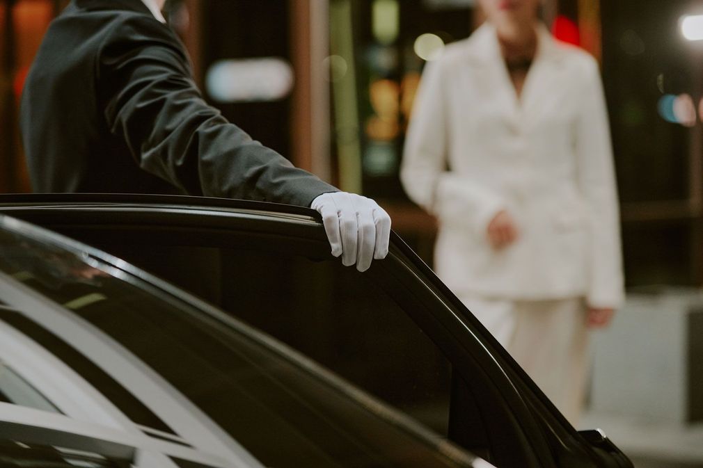 Gloved attendant opening a black car door, with a person in a white suit standing in the background