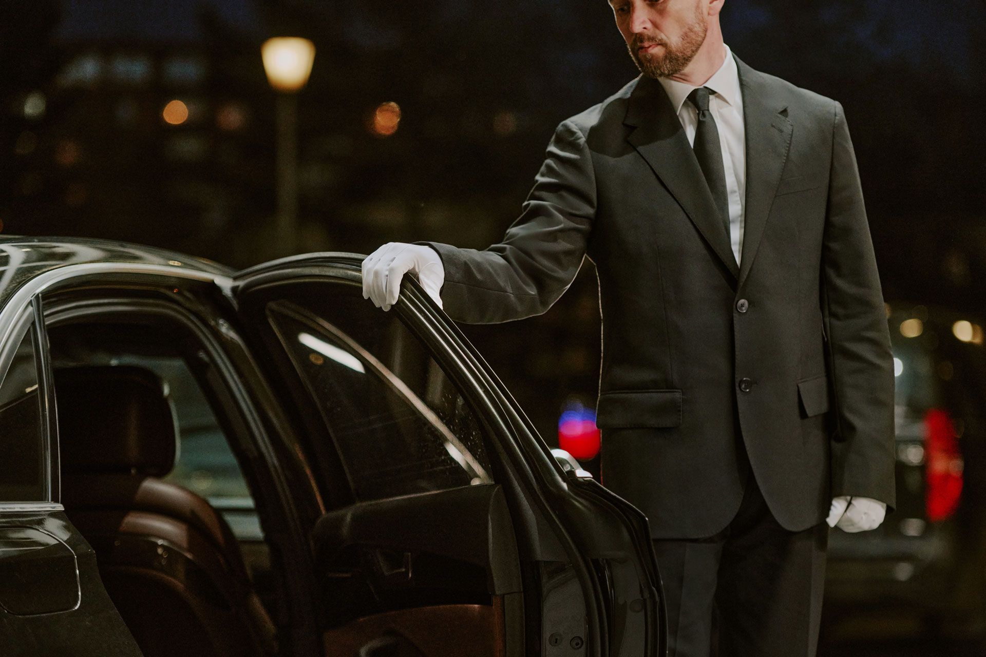 Man in a dark suit and white gloves opening a black car door at night, city lights behind him