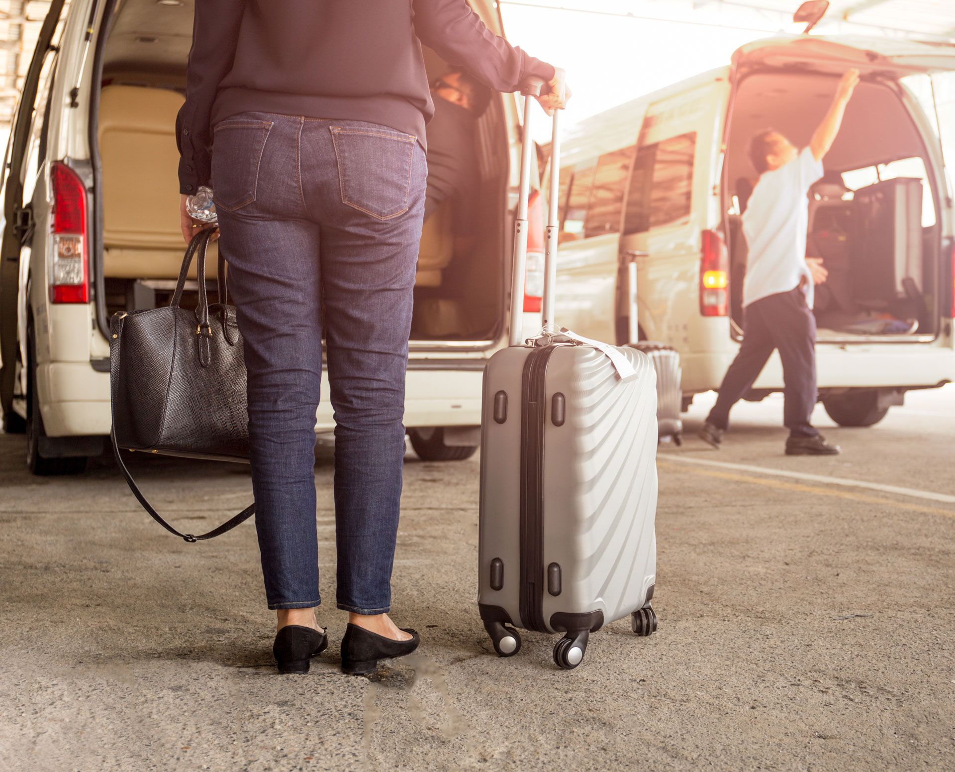 Traveler with silver suitcase beside open van at an airport terminal, other people loading luggage nearby