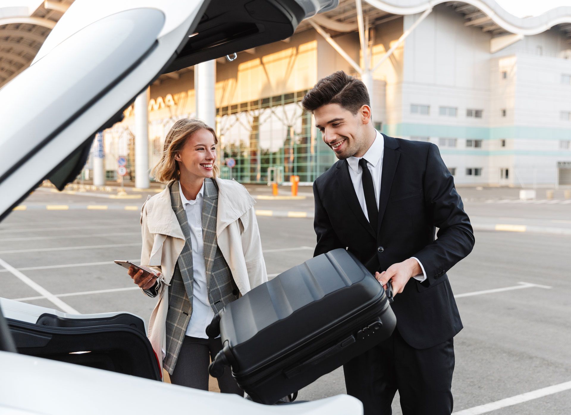 Two people loading a black suitcase into the trunk of a car in a parking lot