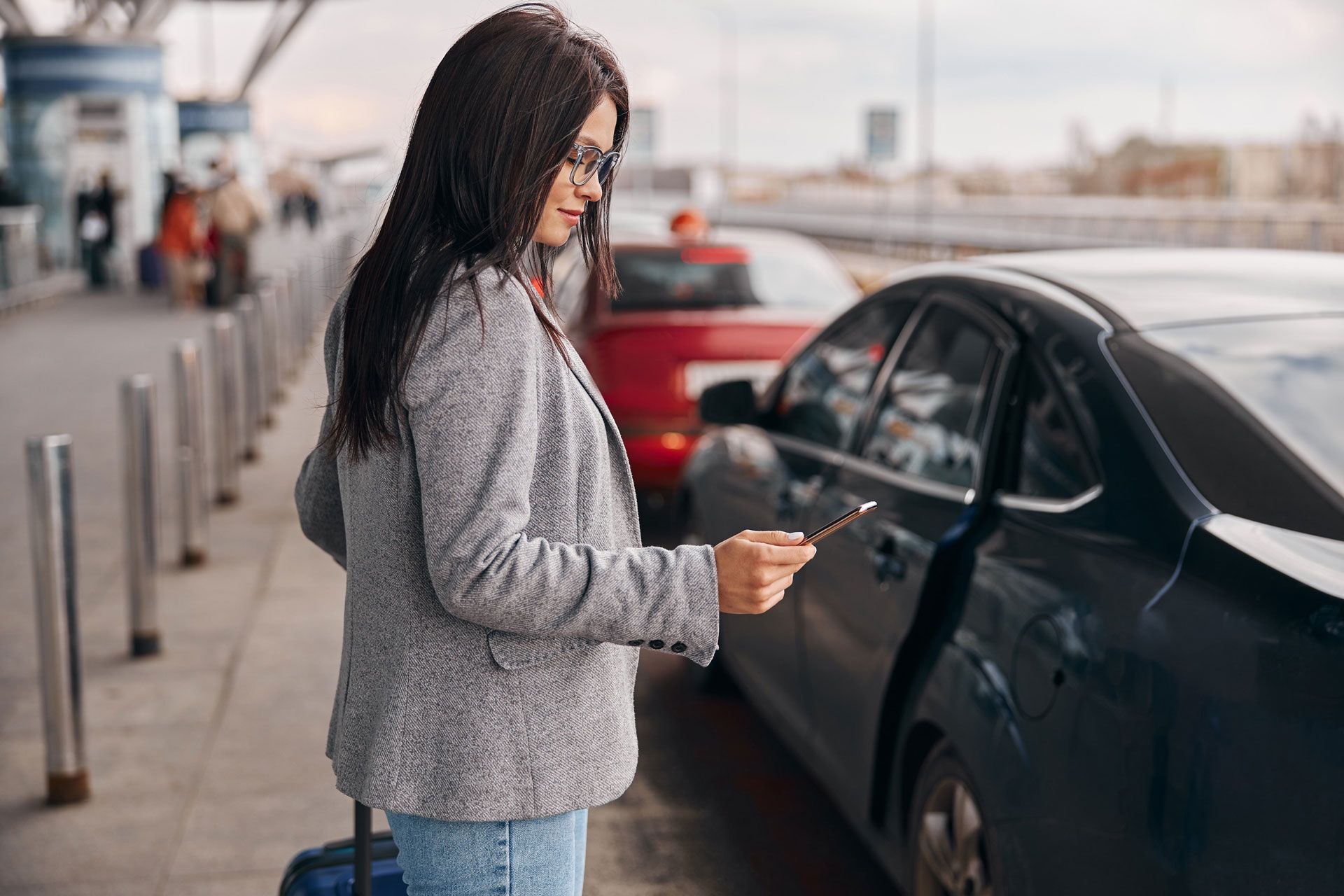 Woman using a smartphone beside a black car at a curbside pickup area