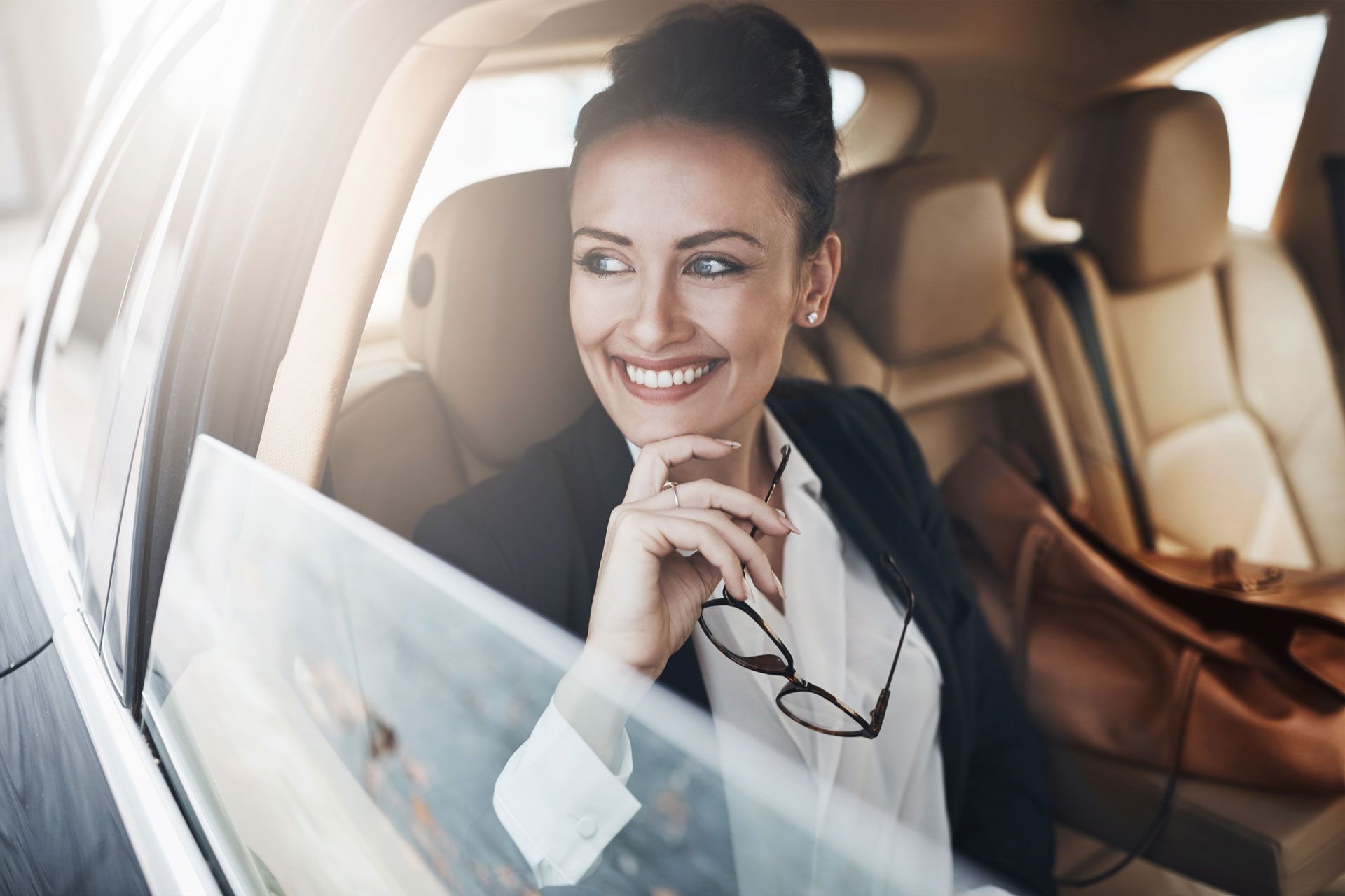 Smiling woman sitting in a car, holding eyeglasses and looking out the window.