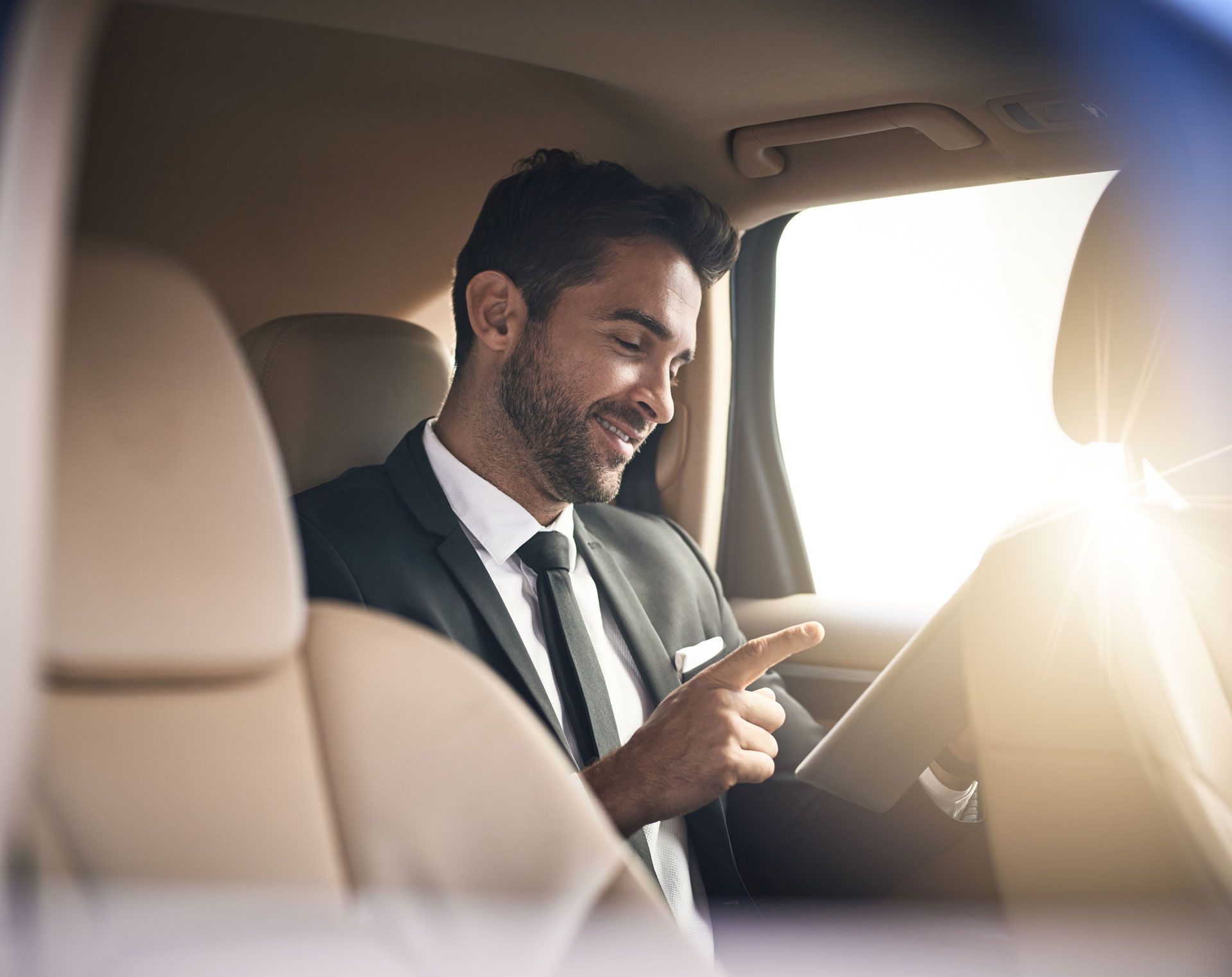 Man in a suit smiling and using a tablet in a car, with bright sunlight through the window