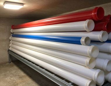 Stacked red, white, and blue cylindrical pipes stored on a metal rack in a warehouse.