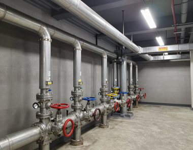 Industrial utility room with large silver pipes, red valves, and gray concrete walls.