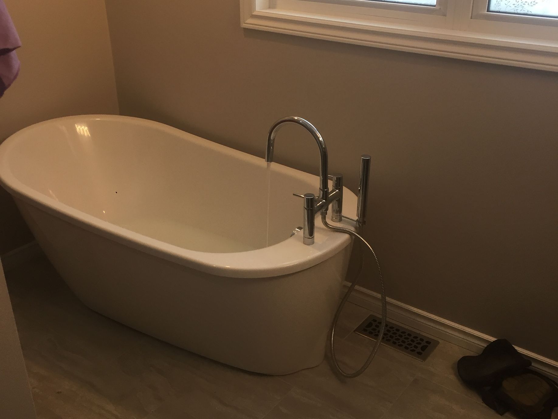 Freestanding white bathtub filling with water in a bathroom; tan walls, window.