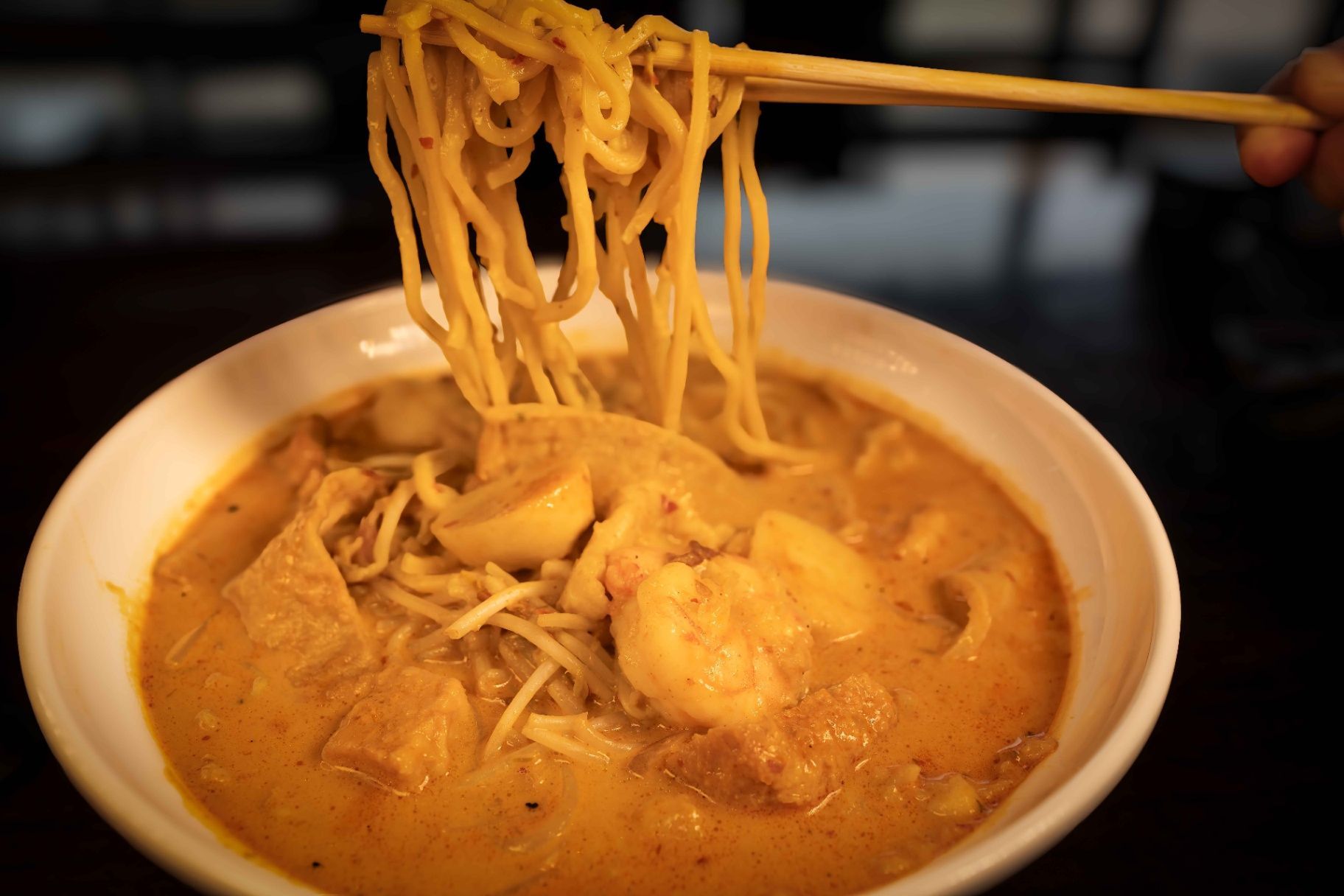 Chopsticks lifting noodles from a bowl of orange soup or curry
