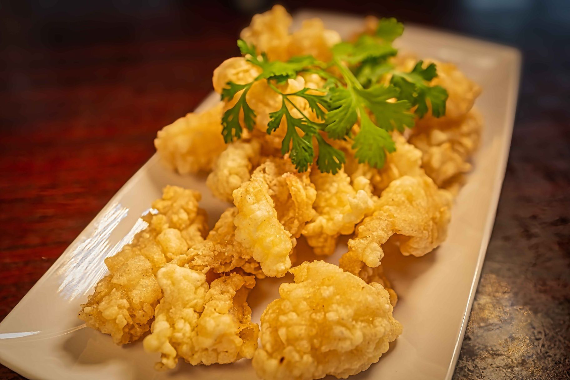 Golden fried chicken bites on a rectangular plate, topped with fresh parsley, on a dark tabletop