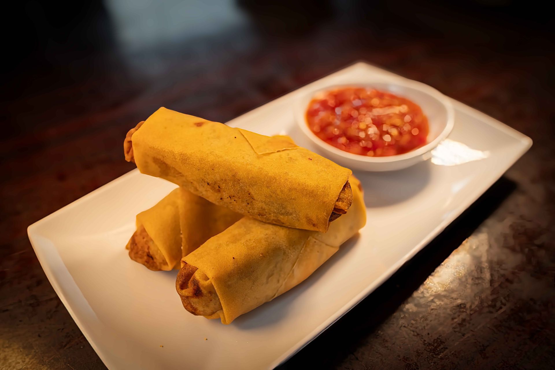 Two rolled tacos on a white plate with a bowl of salsa on a dark table