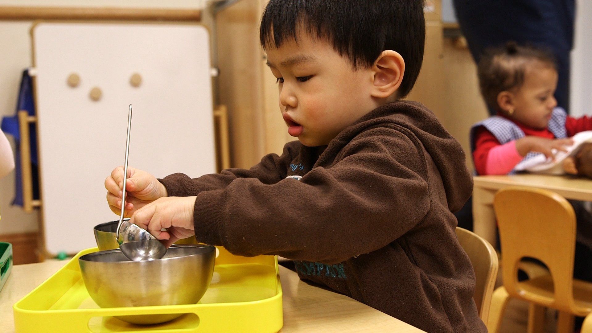Young child stirring a metal bowl with a spoon, indoors at a table with other children.