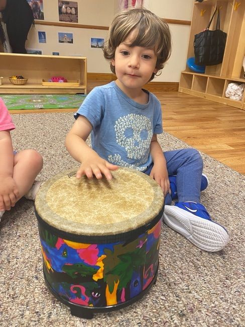 Boy playing a drum, sitting on the floor. He wears a blue shirt with a skull design and leggings. The drum is colorful.