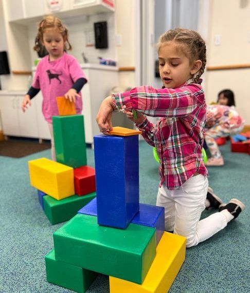 Two young girls playing with colorful building blocks on a blue carpet. One girl is kneeling and placing a block.
