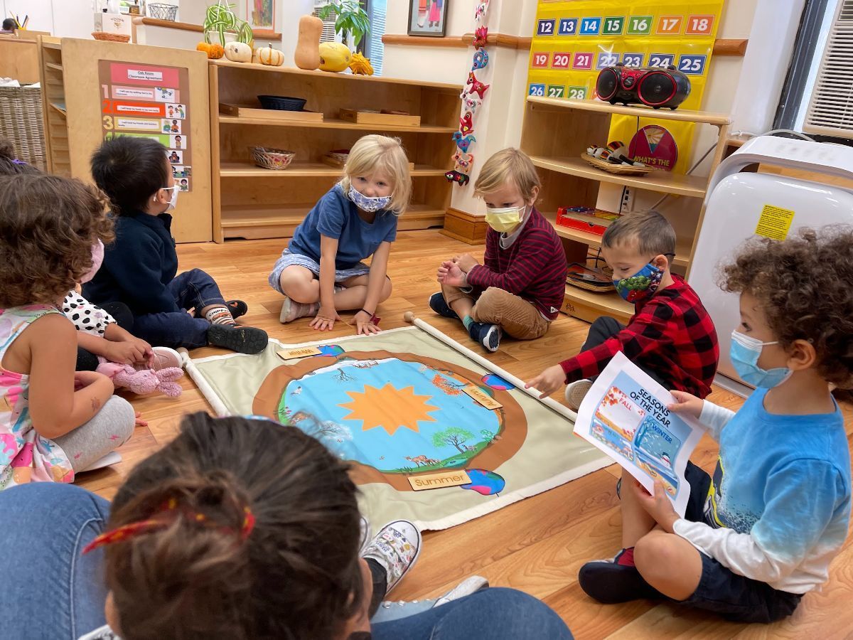 Children wearing masks gathered, listening to a teacher reading a book about a pond.