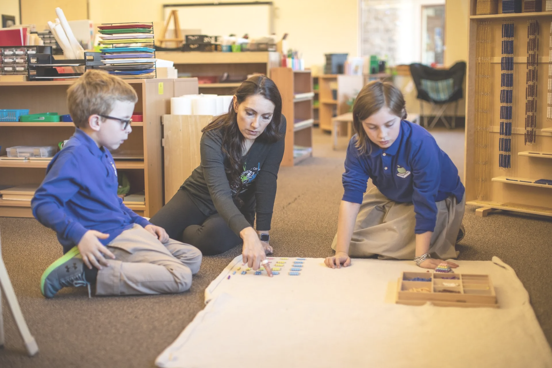Teacher and two students in a classroom. Teacher points at materials on a mat; children watch.