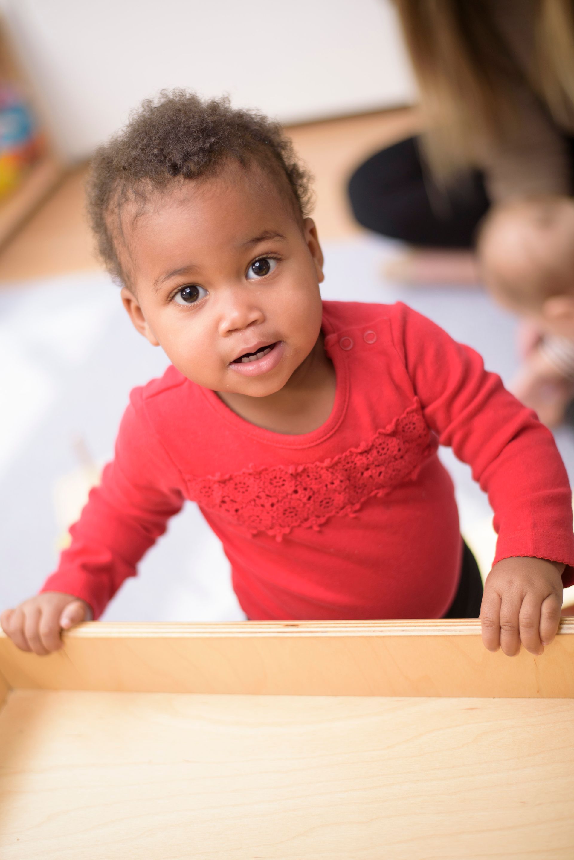 Toddler in a red shirt standing at a table and looking toward the camera