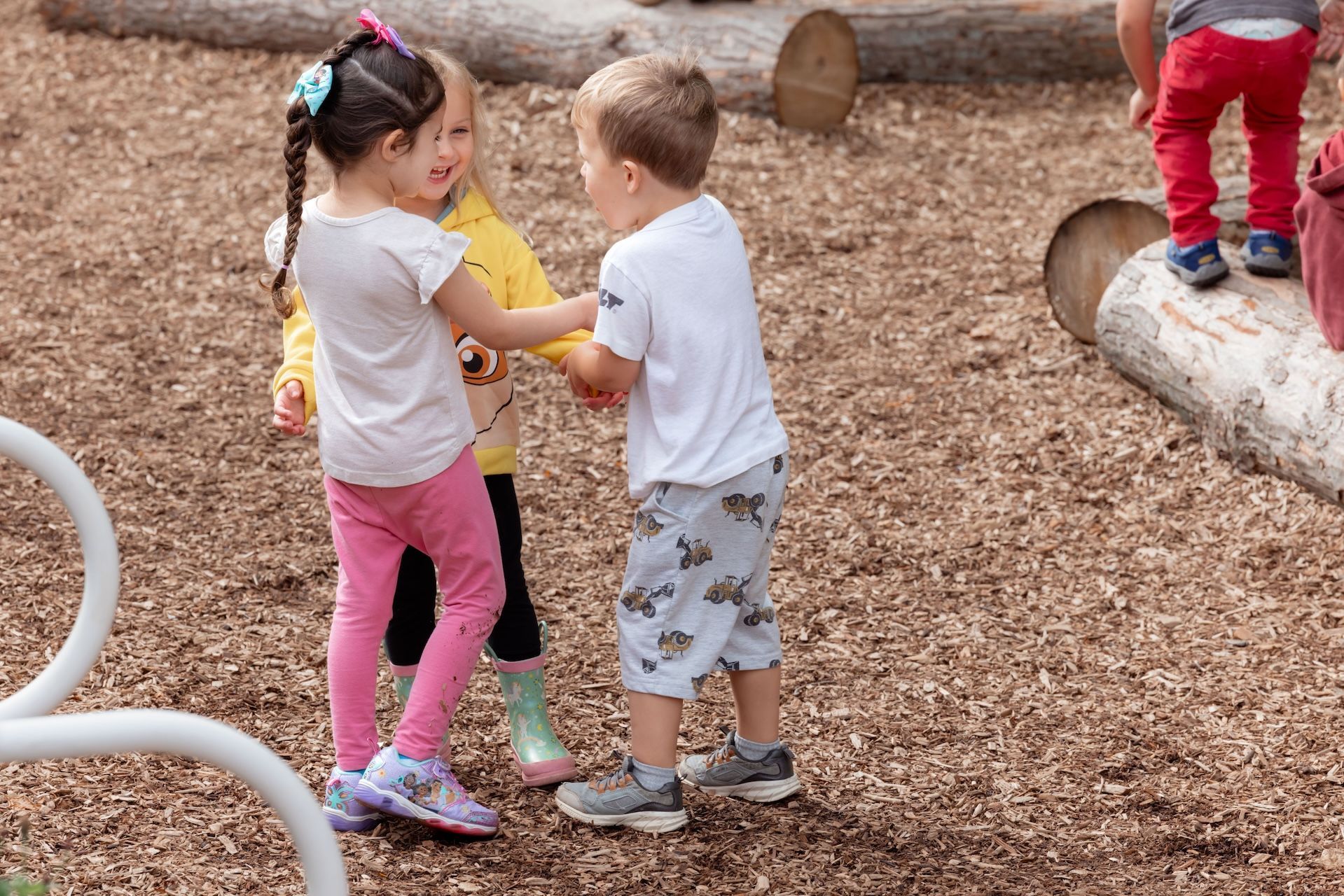 Children playing on a playground. Girl in pink pants, boy in shorts, and girl in yellow shirt holding hands.