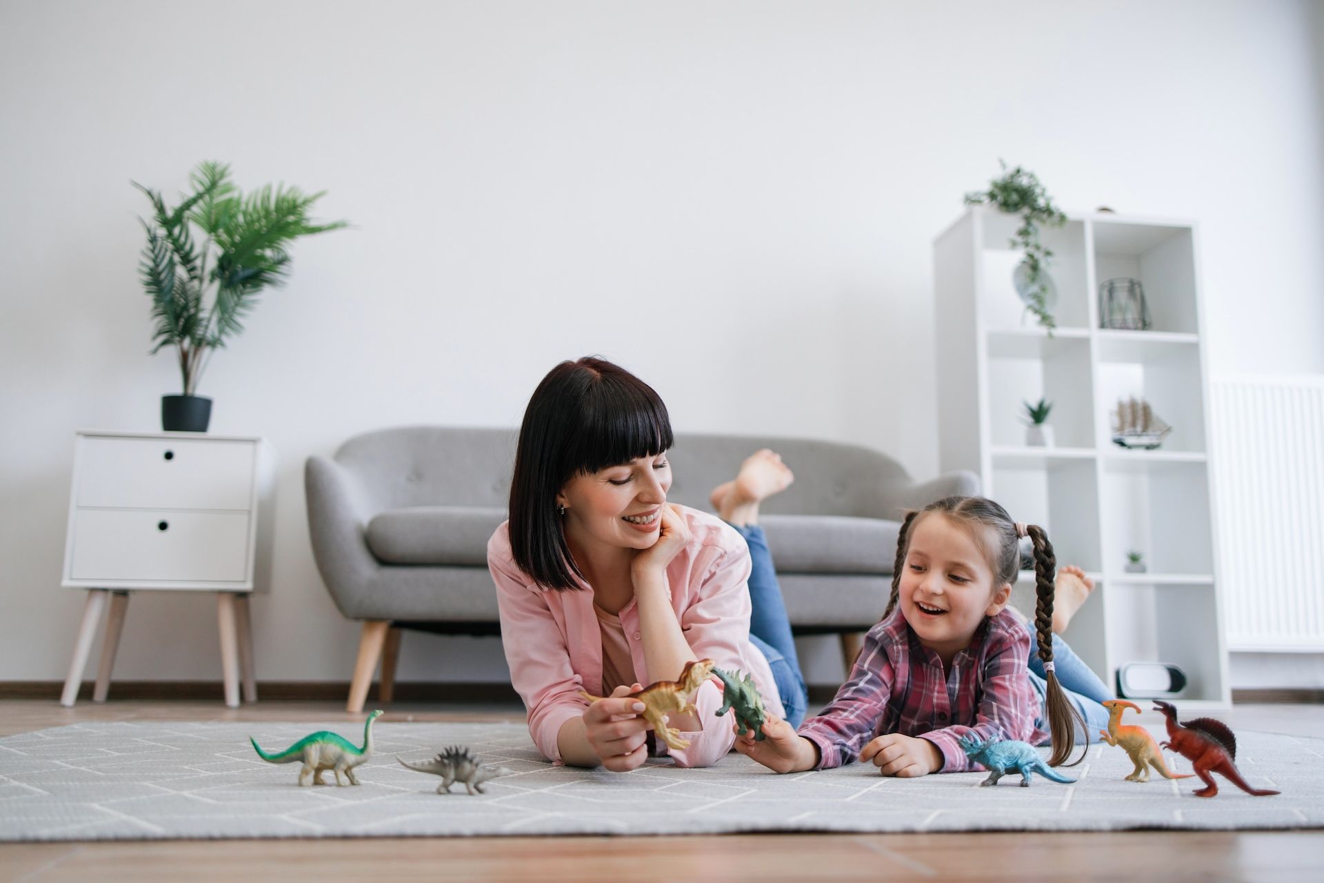 Woman and child playing with toy dinosaurs on a rug in a living room, smiling.