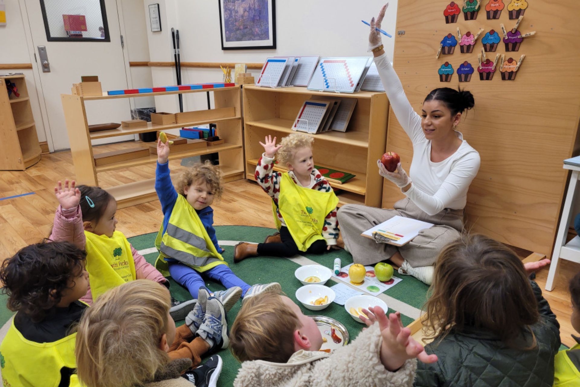 Children and teacher in a classroom circle. Teacher holds an apple; children raise hands.