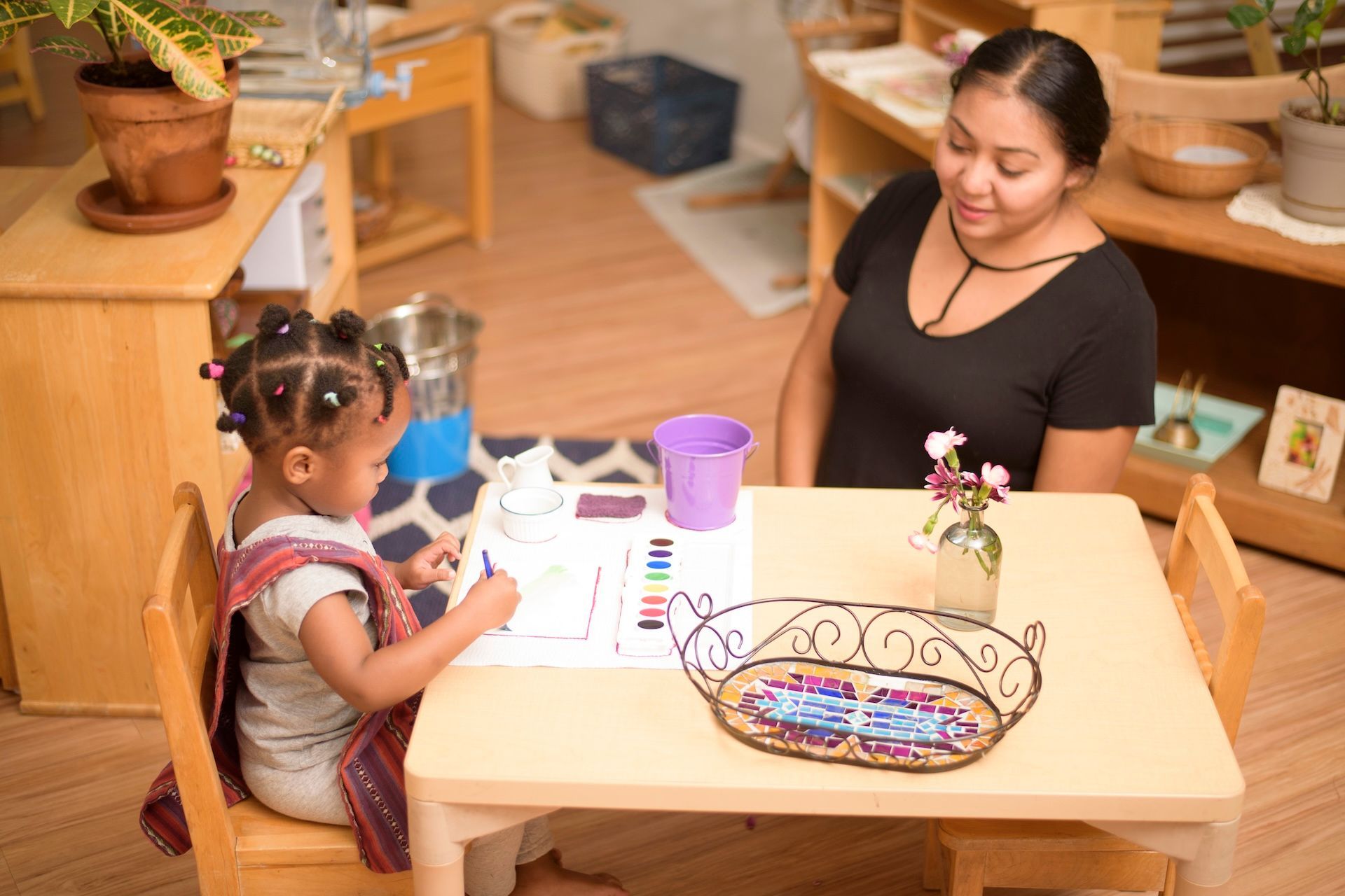 Child and woman sitting at a table coloring in a cozy kitchen with a basket and purple cup