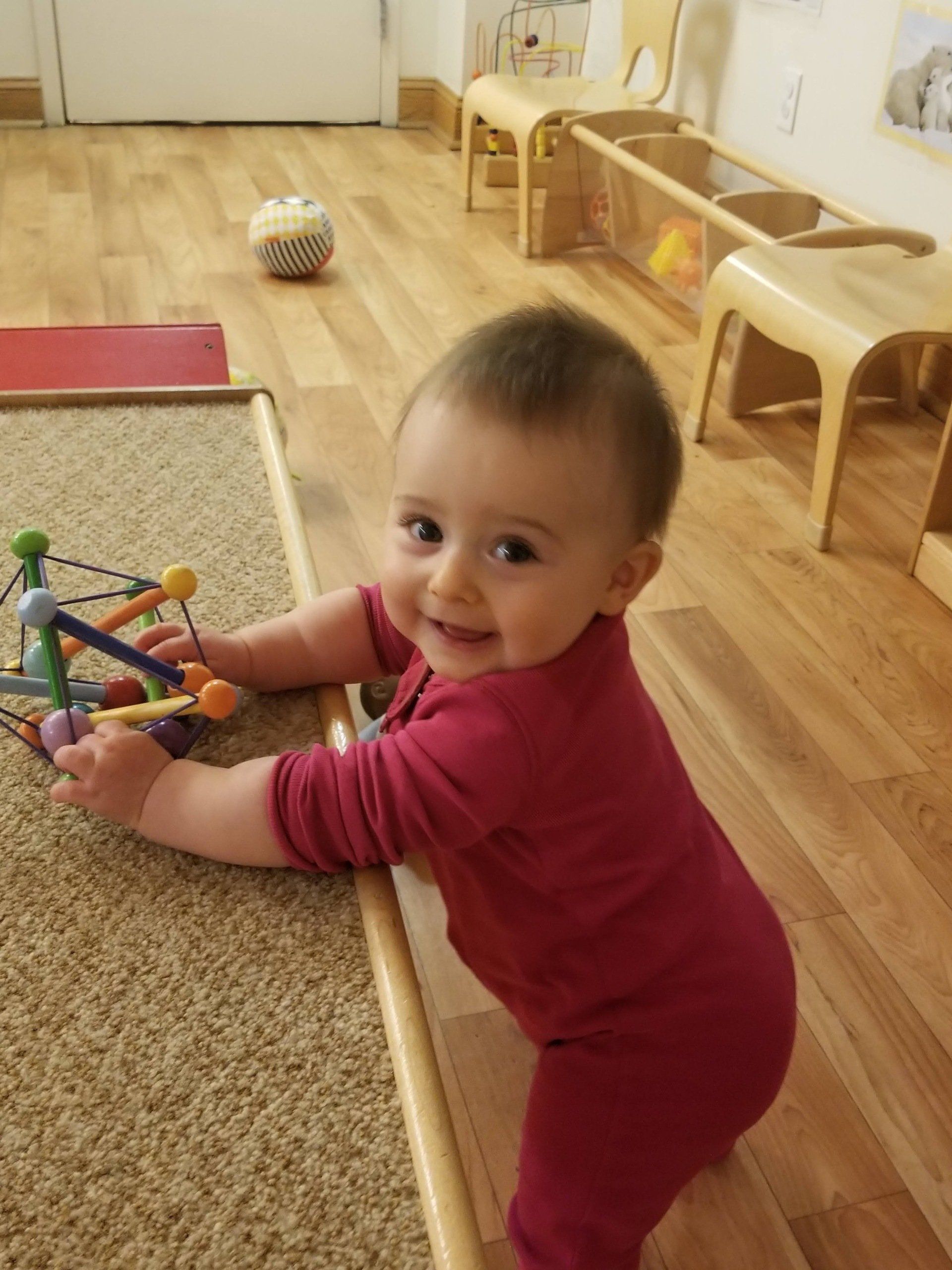 A Montessori infant standing near a toy, wooden floor, and furniture in the background.