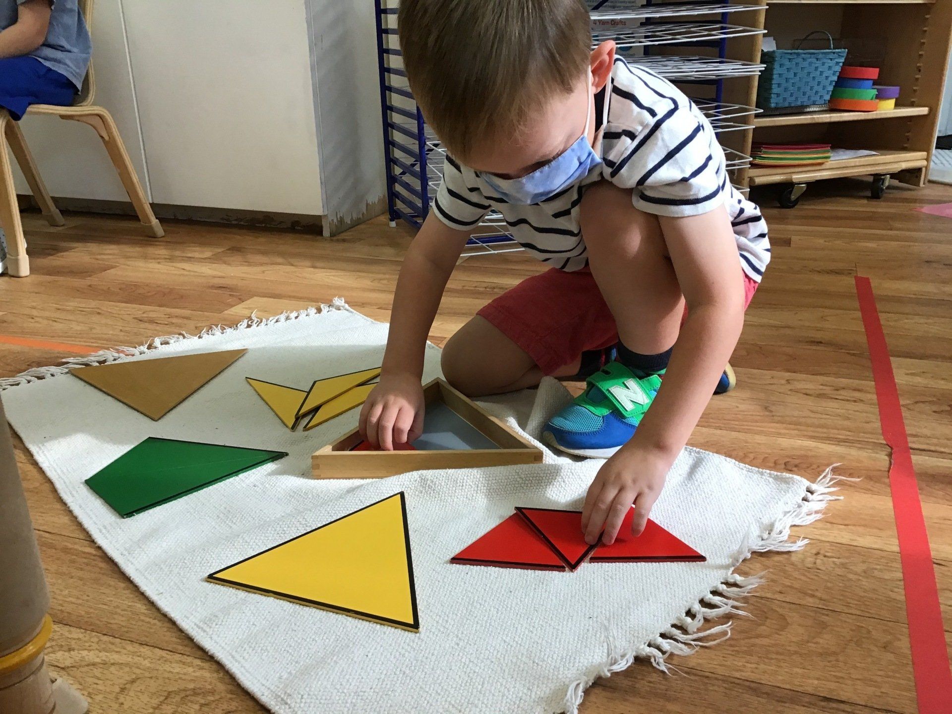 Boy wearing a mask plays with colorful geometric shapes on a rug indoors.