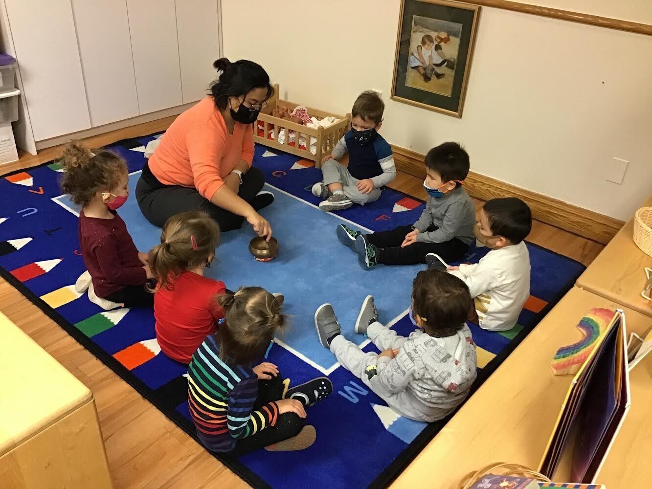 A teacher with mask and students in a circle on rug; they are focused on an object.