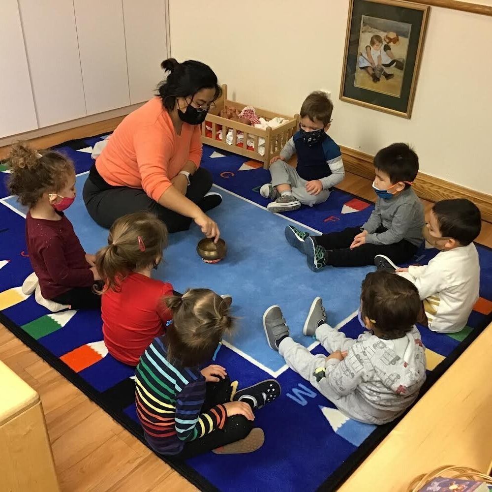 A group of children are sitting in a circle on a rug with a montessori guide playing a bell.
