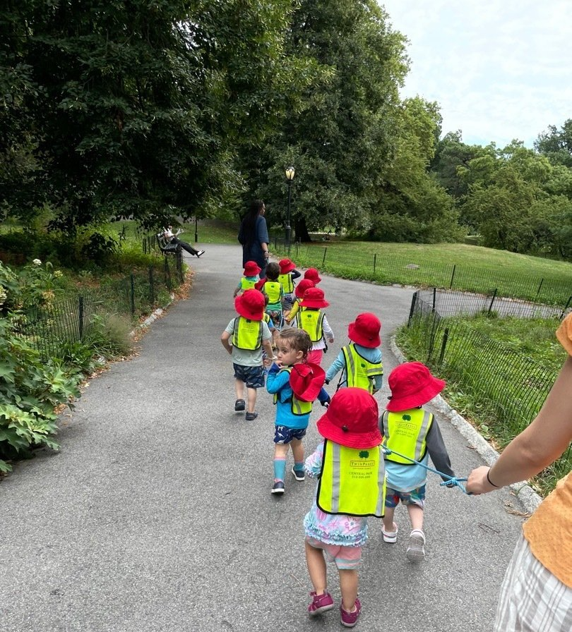 Montessori childs wearing red hats and yellow vests walk in a park, holding hands. Trees and green grass are in the background.