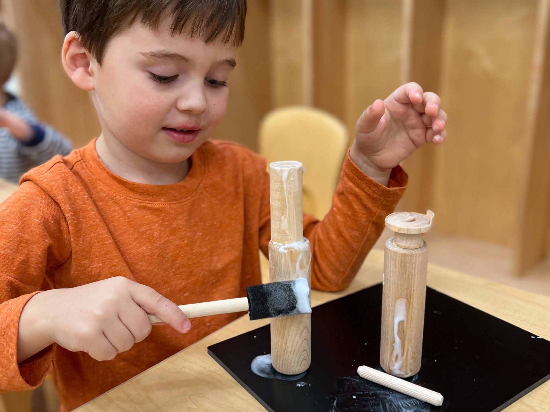 Boy in orange shirt gluing wooden objects at a table.