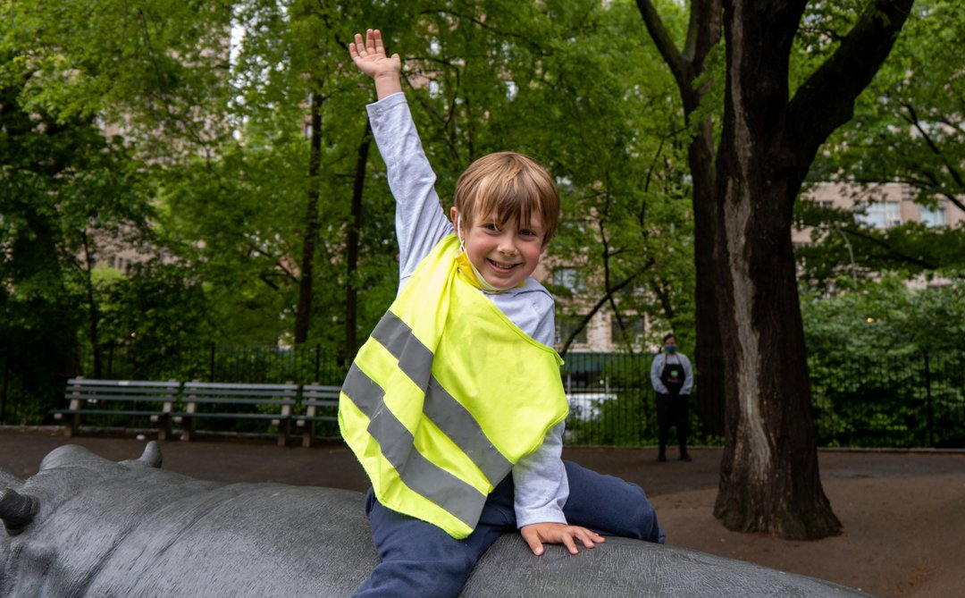 Boy in a yellow safety vest waves from atop a sculpture in a park.