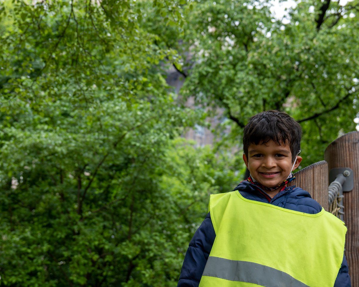 Smiling boy wearing a yellow safety vest stands near trees and a wooden structure.